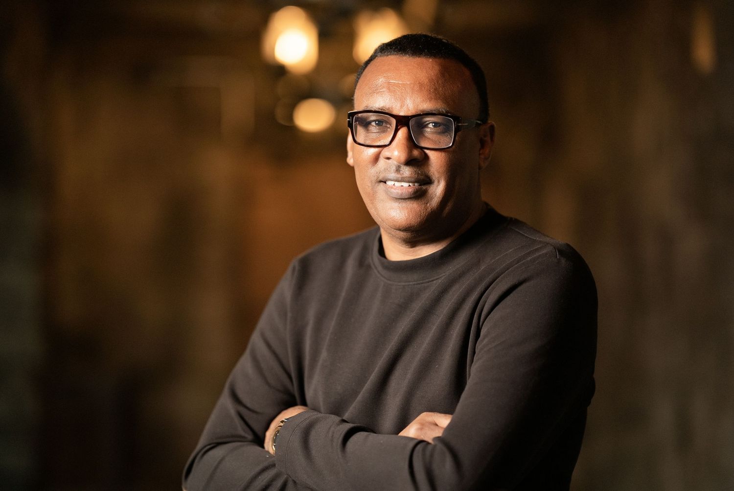 A smiling man with glasses and a dark sweater stands with arms crossed against a warm, dimly lit background.