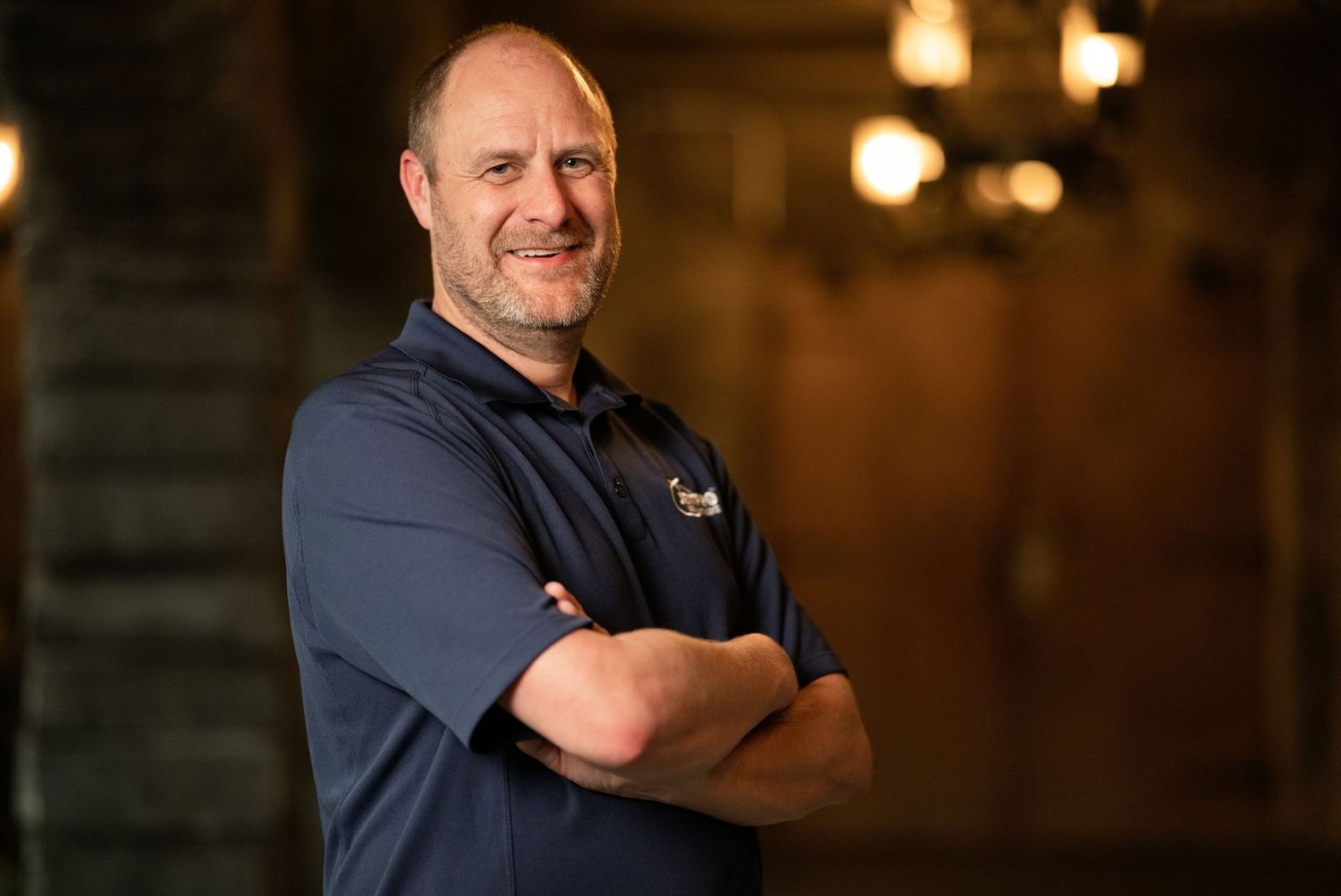 Man in navy polo shirt standing with arms crossed, smiling confidently in a warmly lit rustic interior.