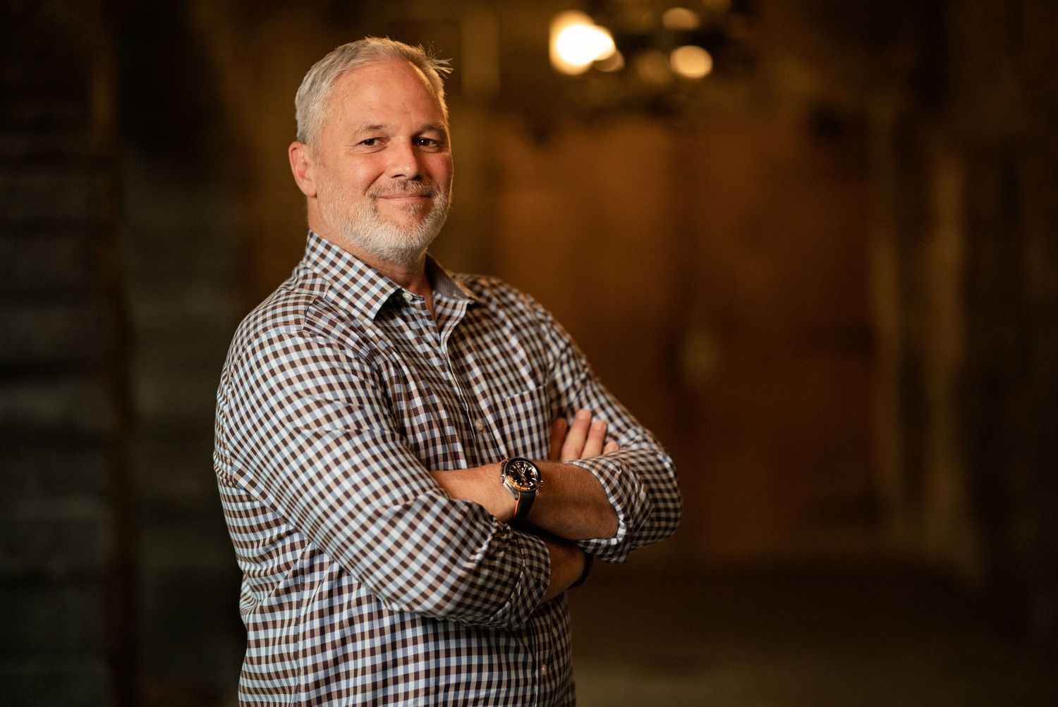 Smiling middle-aged man with gray hair, arms crossed, wearing a plaid shirt, posing confidently in a warmly lit venue.