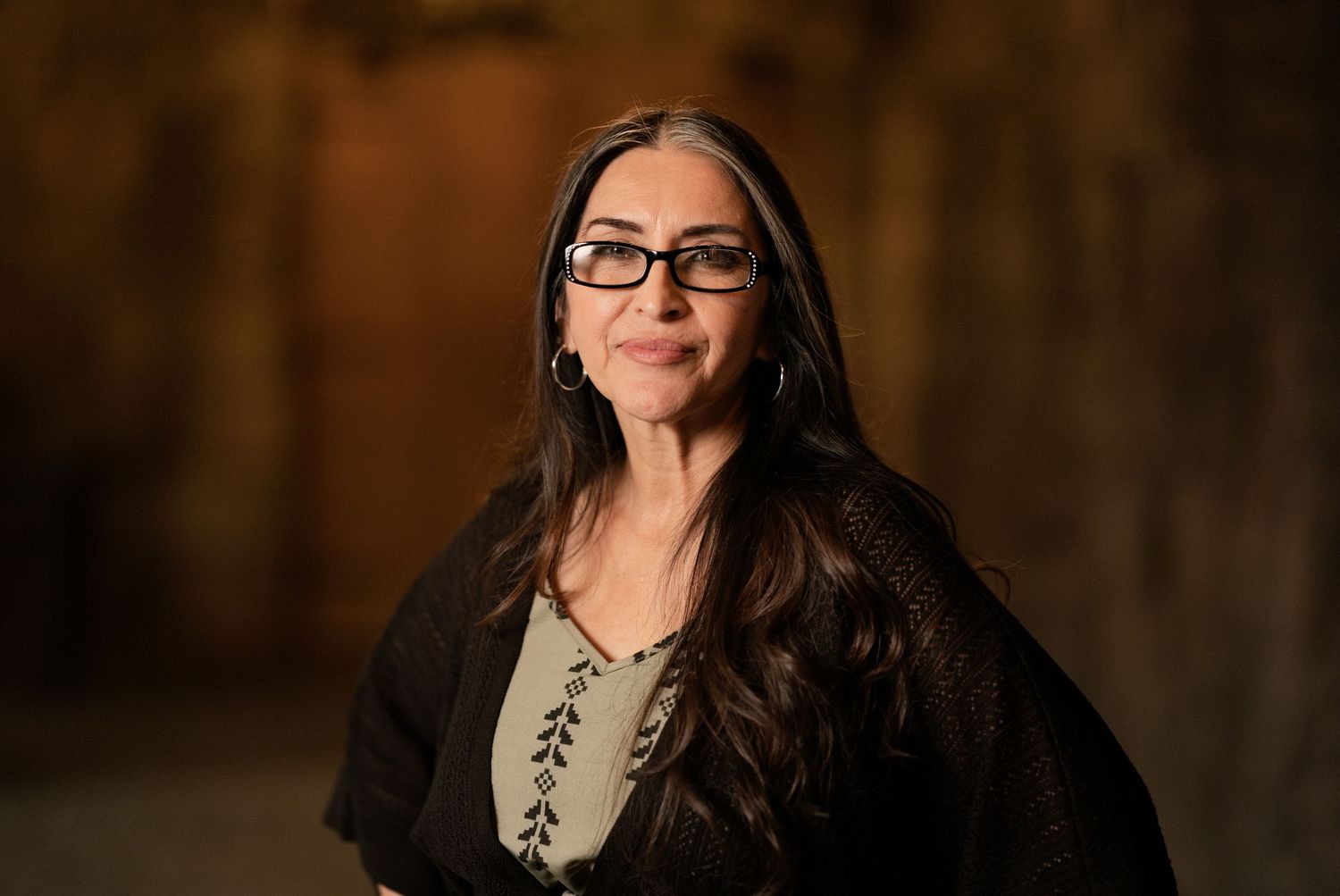 Woman with long dark hair and black glasses wearing a dark shawl, posing confidently in warm ambient lighting.
