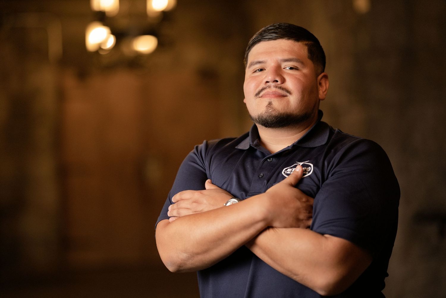 Confident young man in navy polo shirt stands with arms crossed in dimly lit stone venue.