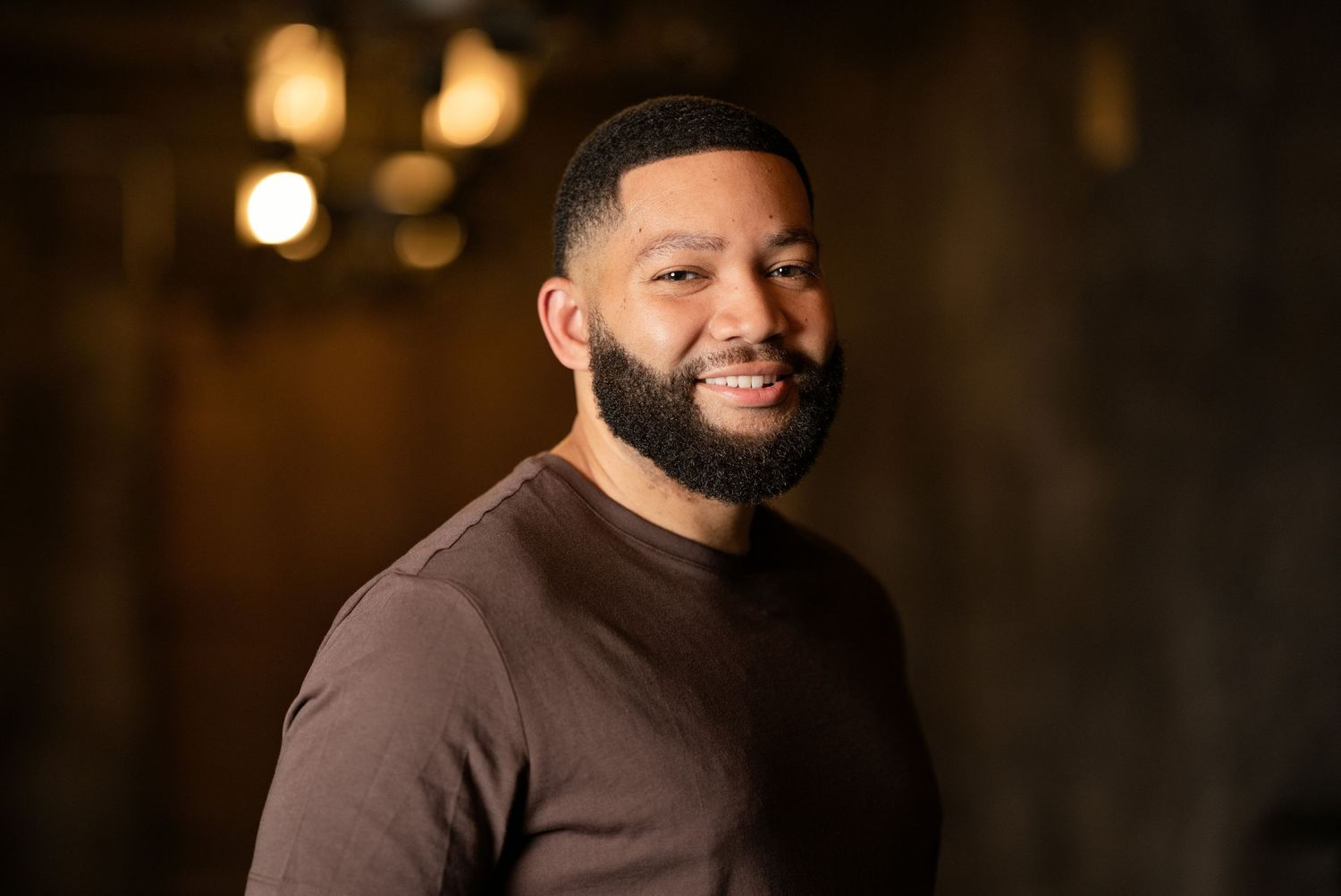 Smiling Black man with beard wearing brown t-shirt poses confidently in warmly lit indoor setting.
