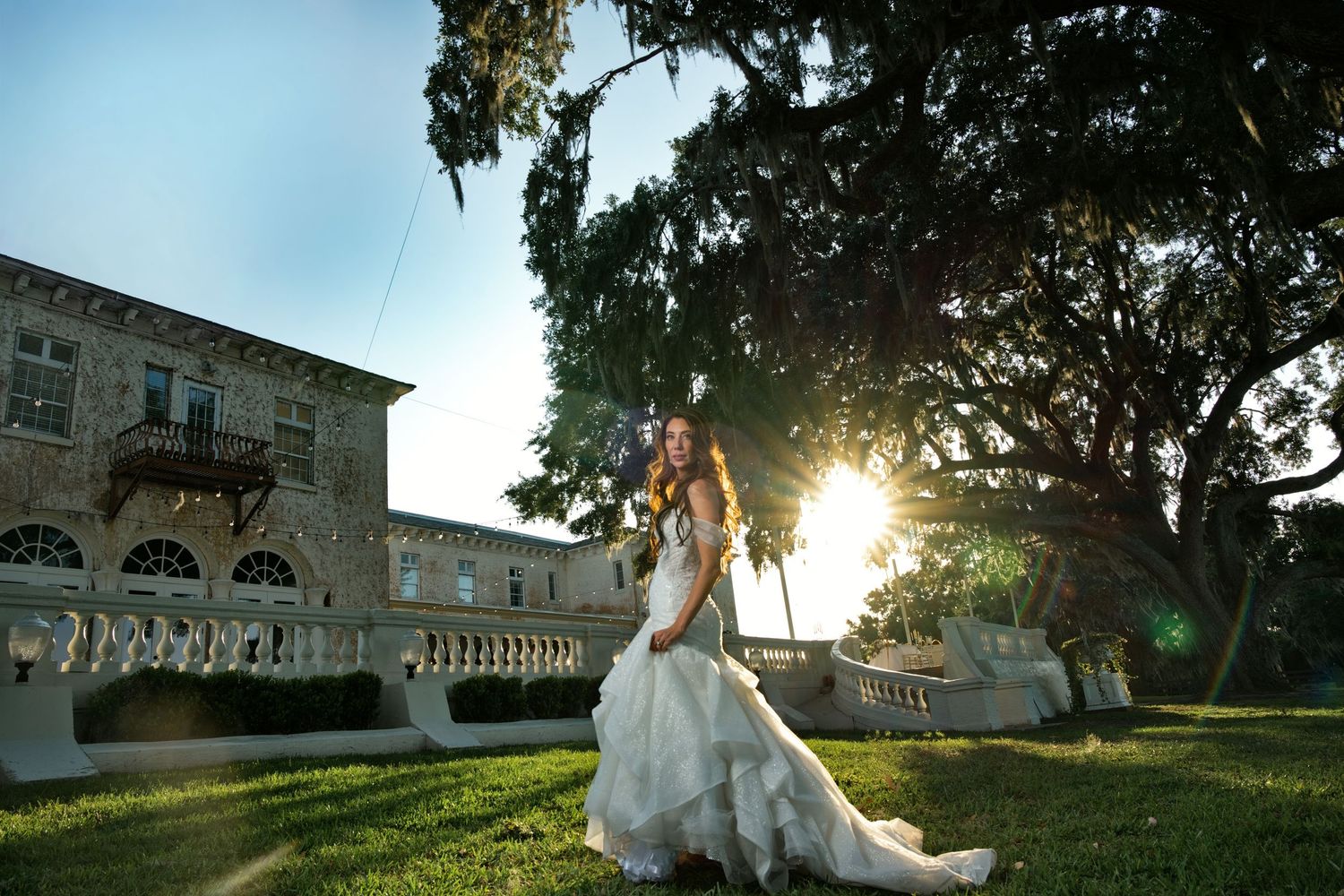 Bride in elegant white gown poses on sunlit lawn before historic mansion with sprawling oak trees.