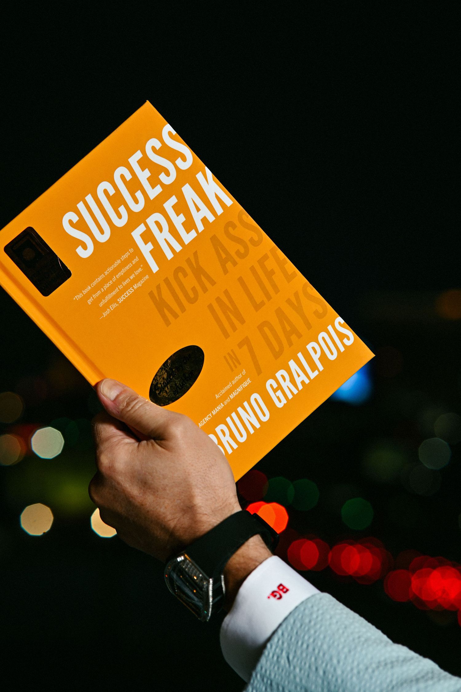 Hand holding orange Success Freak book by Bruno Gralpois against a dark bokeh city light background at night.