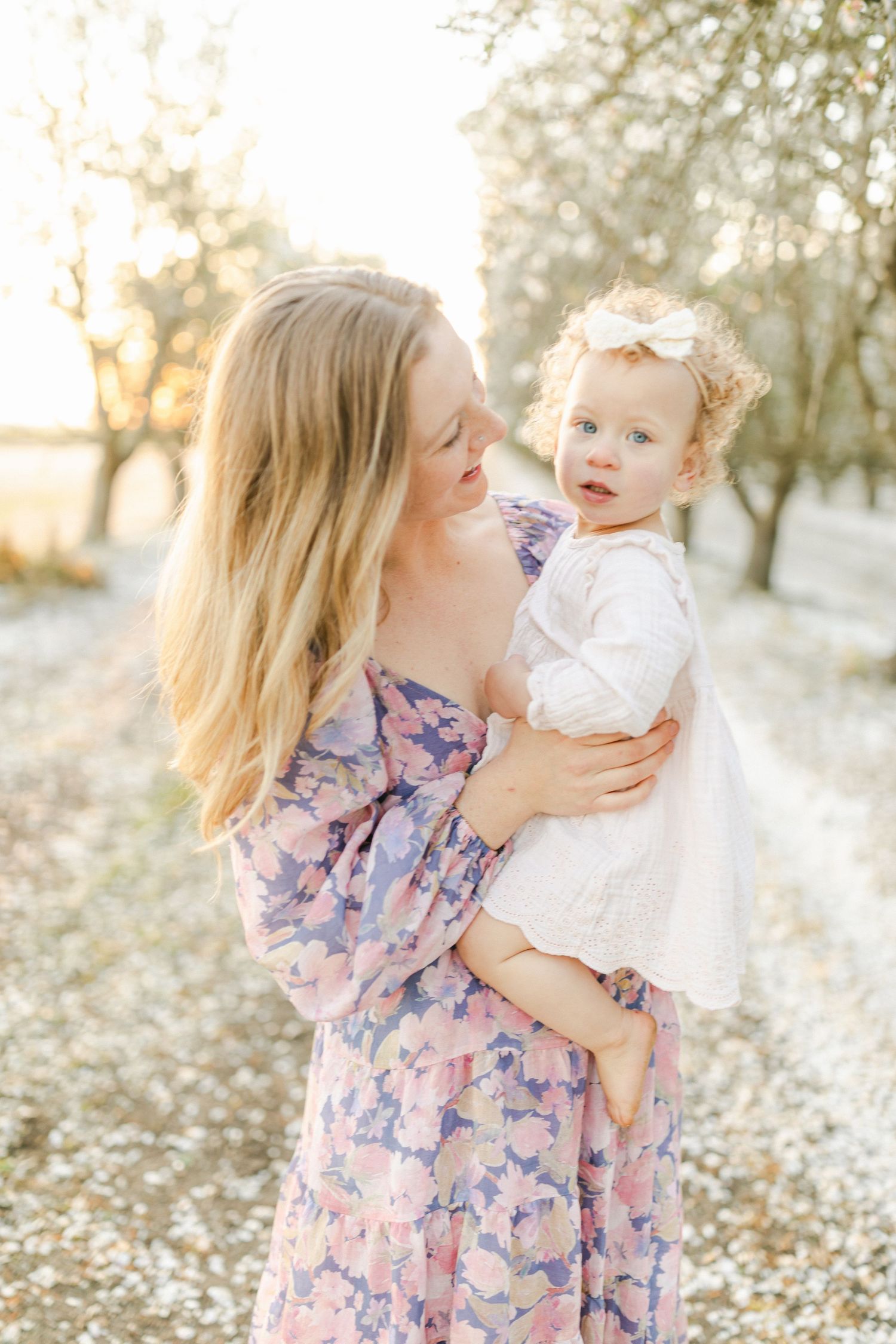 A mother in a floral dress holds her baby girl outdoors among blossoming trees in golden sunlight.