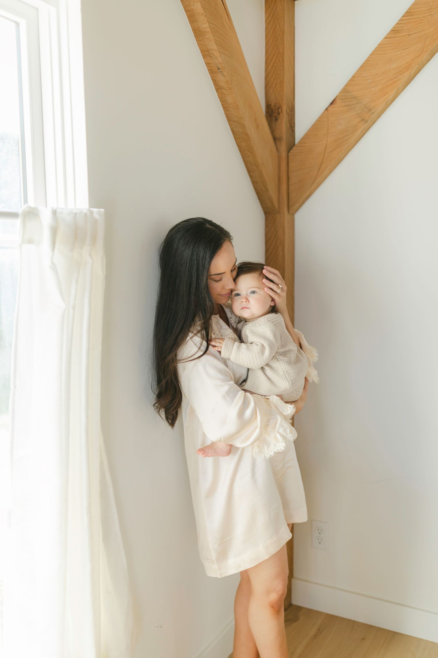 A mother in a cream robe tenderly holds her baby near a bright window with wooden beam accents in a serene white room.