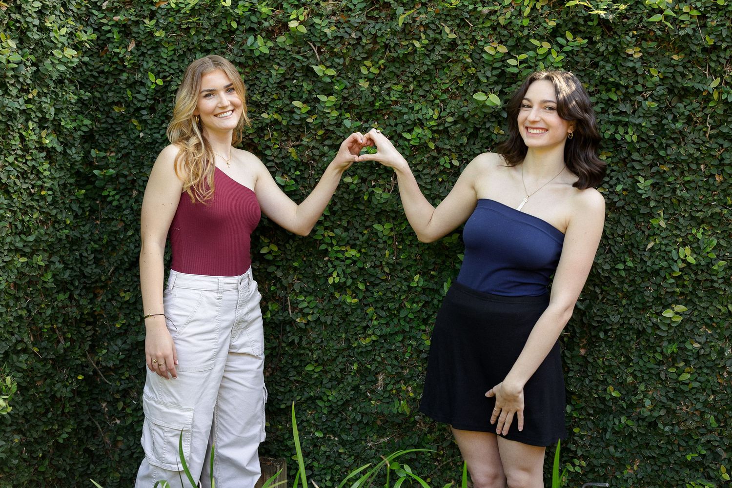 Two people in casual summer outfits pose and laugh together against a green hedge background.