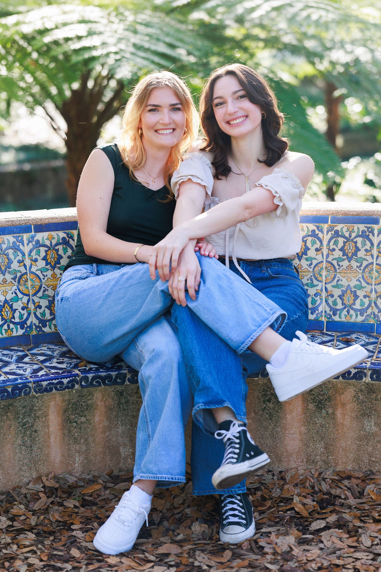 Two friends in blue jeans sit together on a decorative tiled wall surrounded by autumn leaves and greenery.