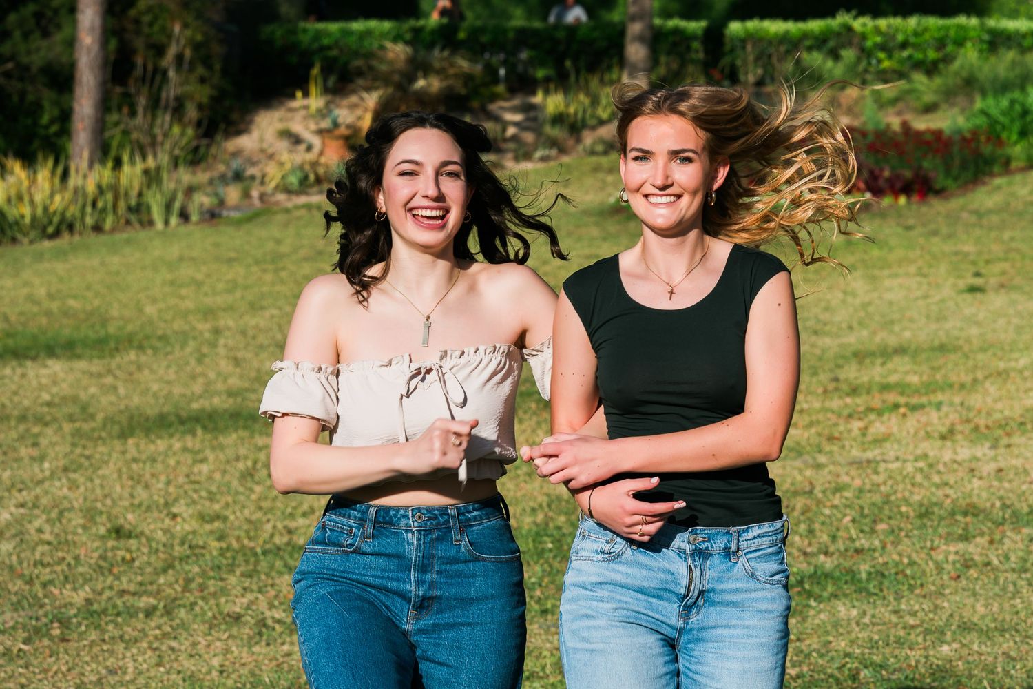 Two people in casual denim and black outfits sit together laughing on grass in a sunny outdoor setting.