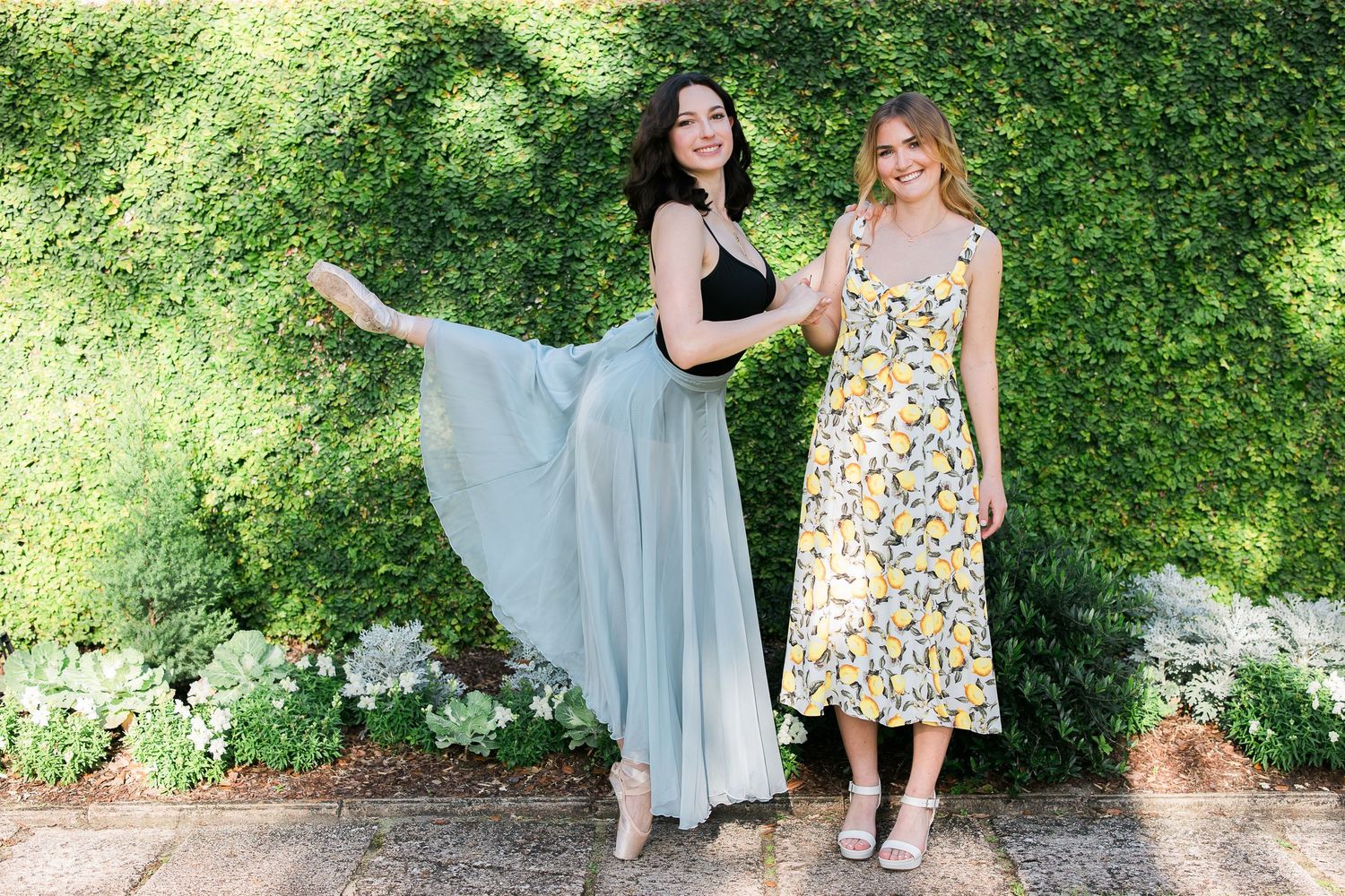 Two people in summer dresses pose together against a lush green garden wall on a sunny day.