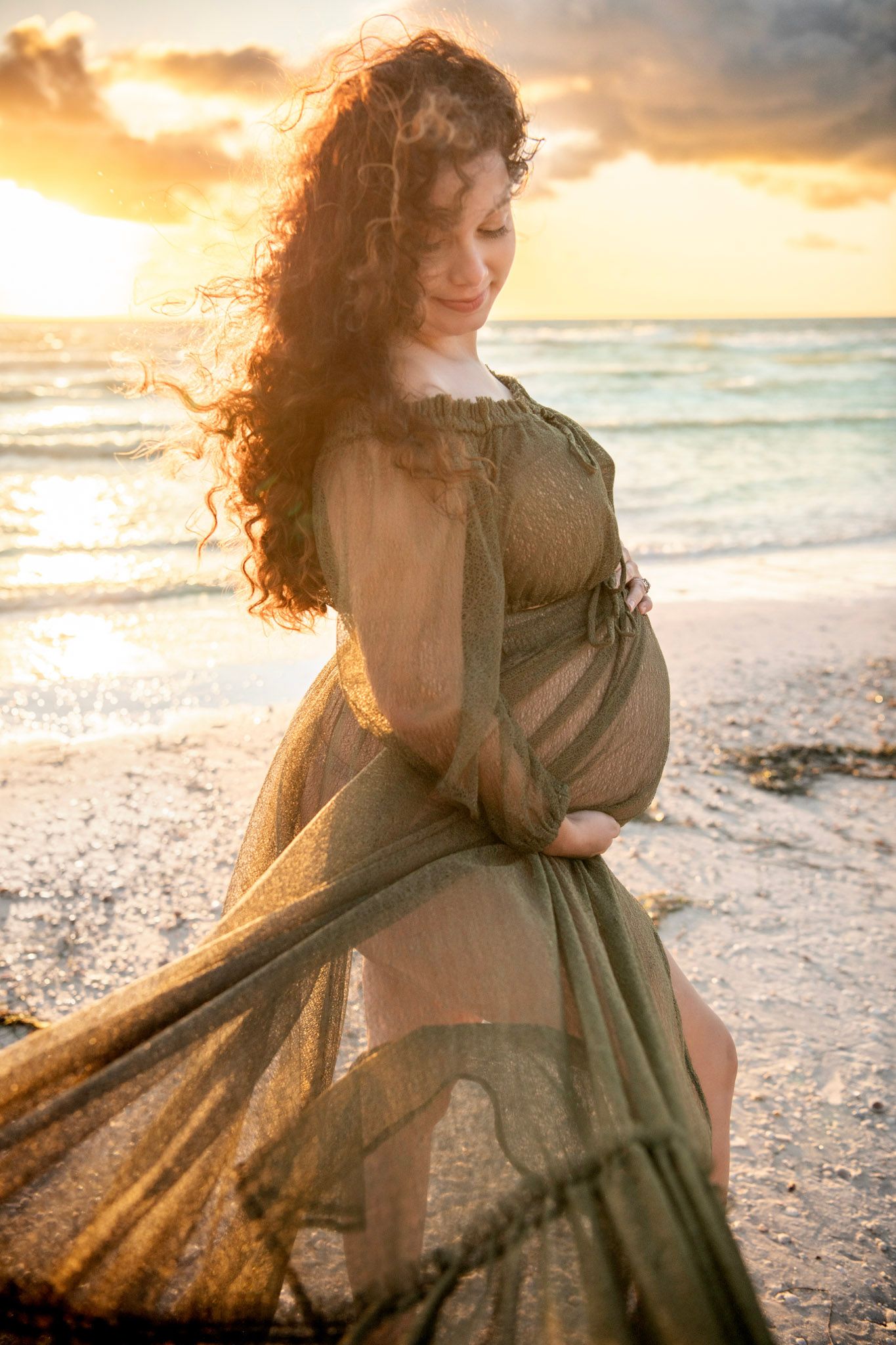 Curly-haired mother-to-be cradles her baby bump on St. Pete Beach, framed by soft waves and glowing sunset light.