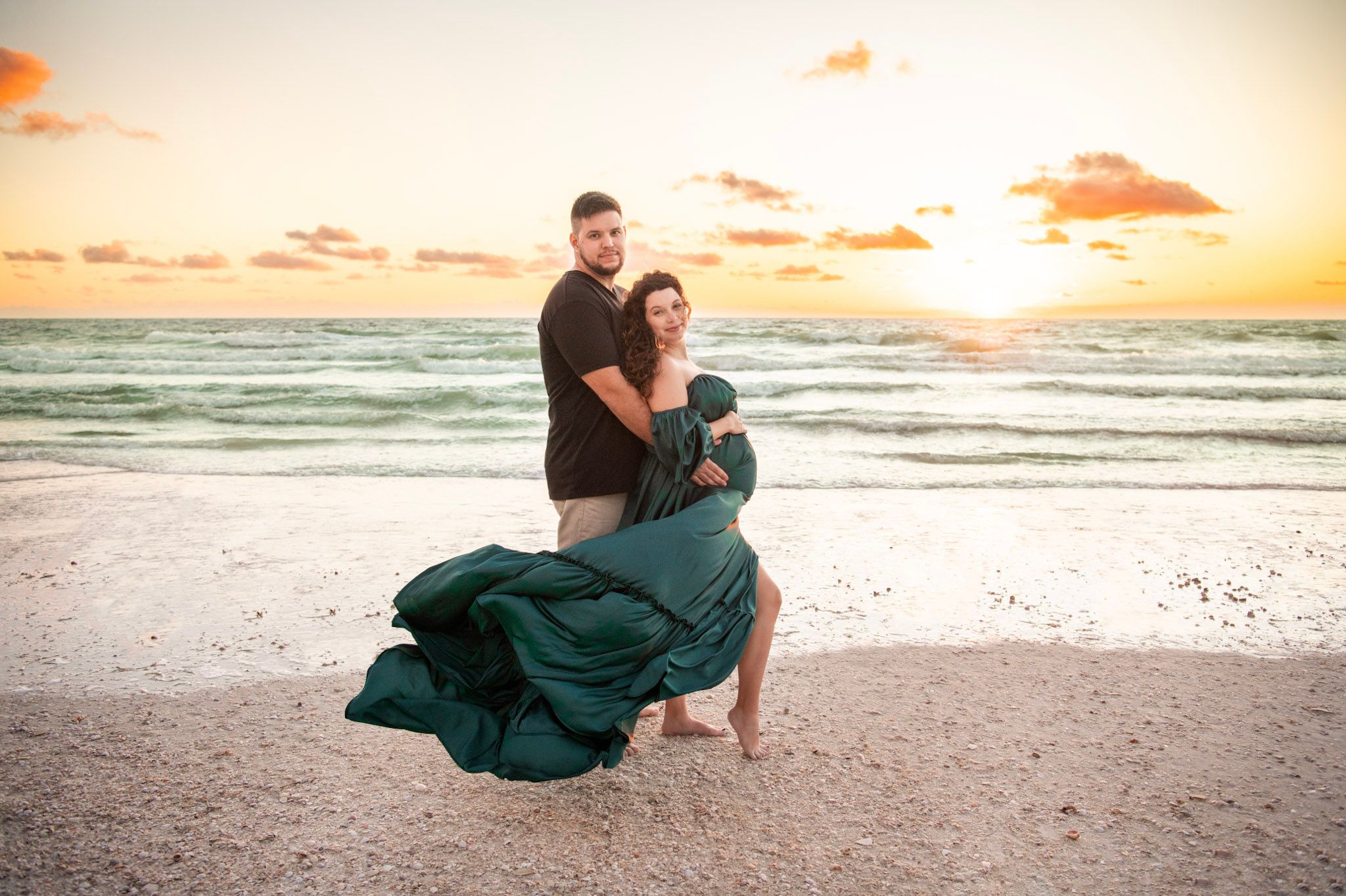 Expecting parents stand together on St. Pete Beach as a dramatic flowing gown billows across the sand at sunset.