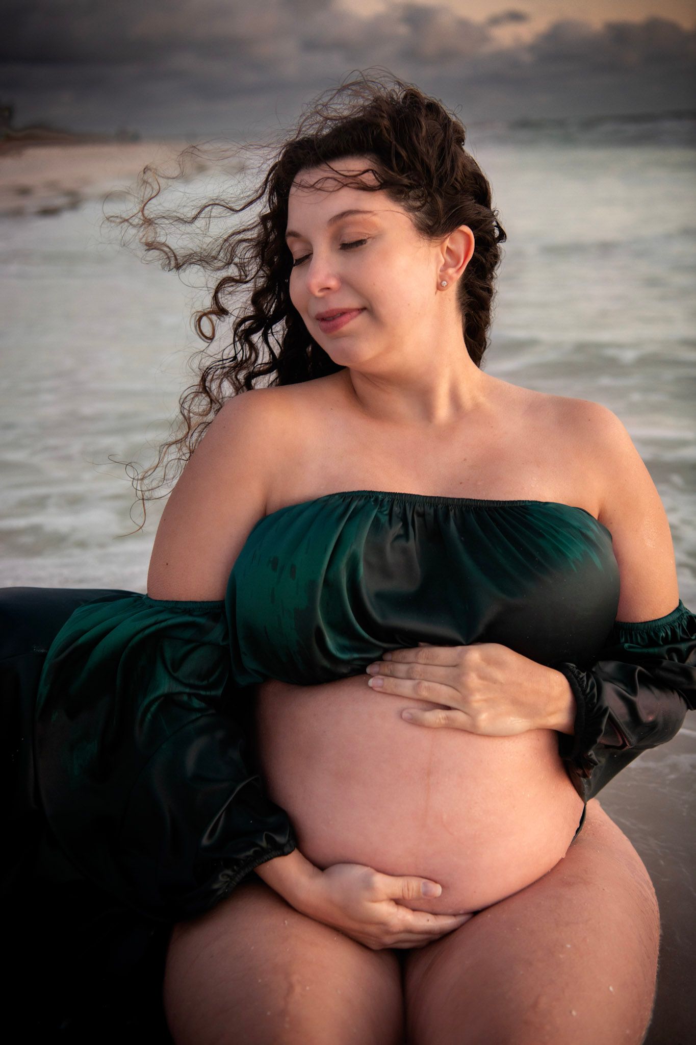 Moody maternity portrait on the shoreline in Dunedin, FL, featuring an expectant mother in a flowing gown beneath dramatic coastal skies.