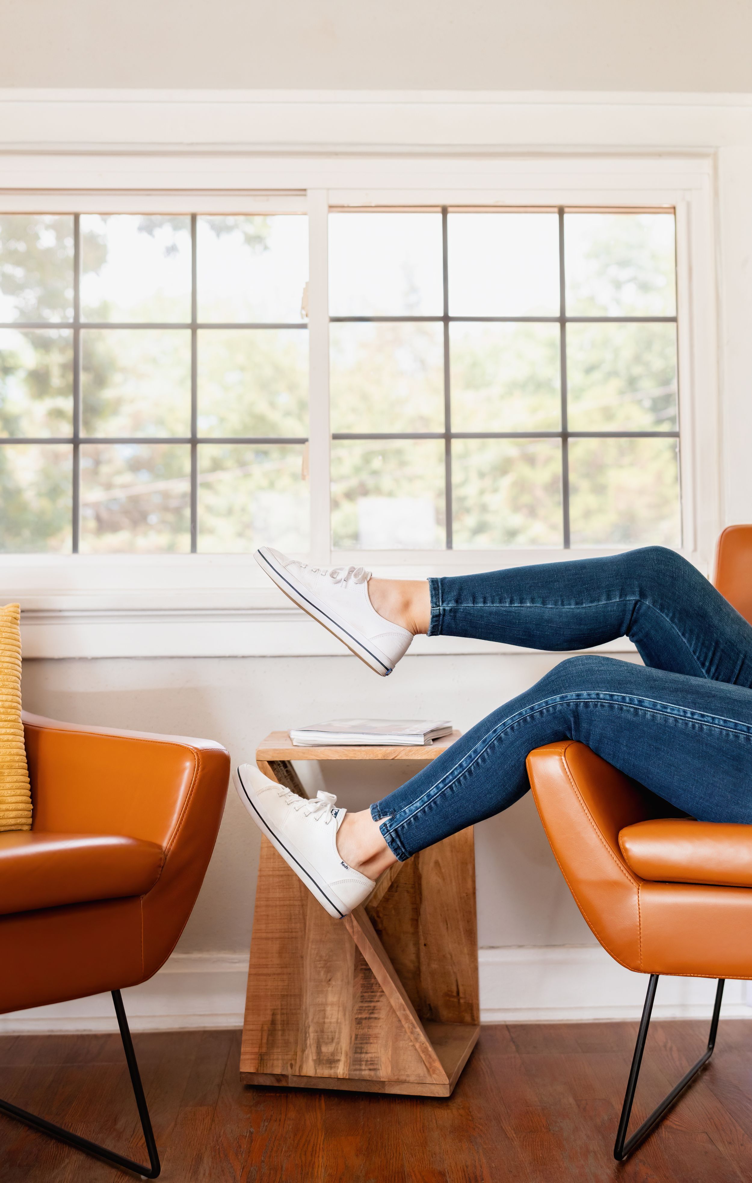 Person relaxing in orange leather chair with feet up on wooden coffee table, wearing jeans and white sneakers near bright window.