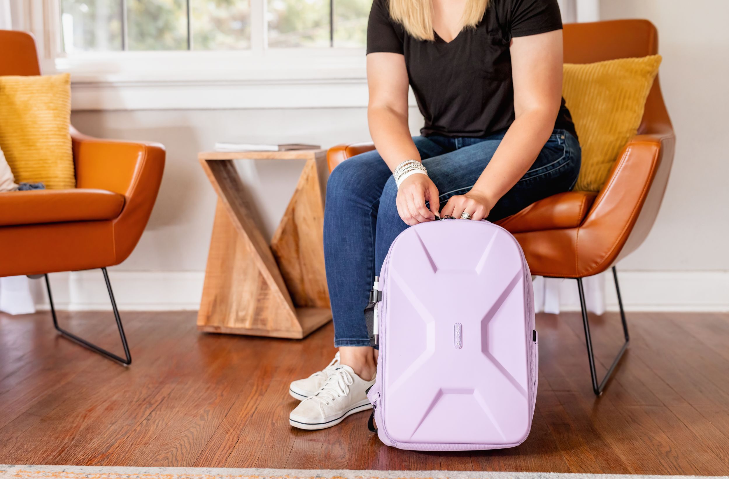 Woman sitting on a chair zipping a lavender hard-shell backpack in a modern living room.