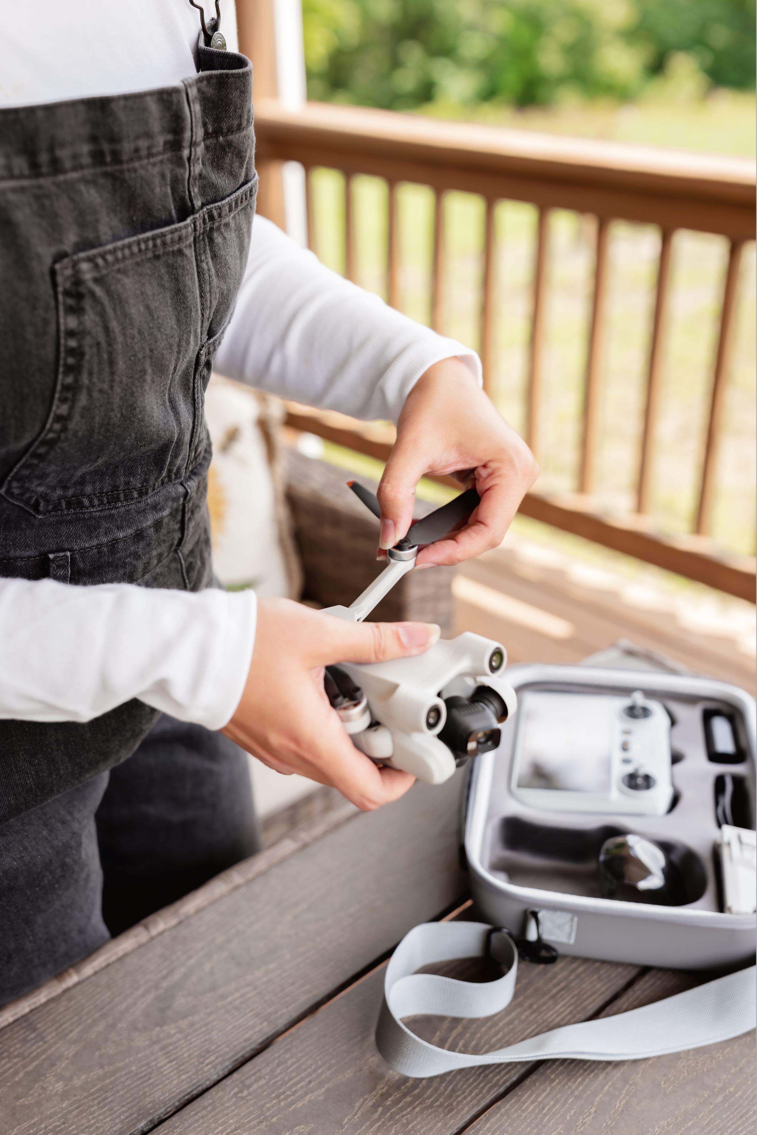 Person in denim overalls assembling a drone outdoors, with a carrying case and remote controller on a wooden deck.