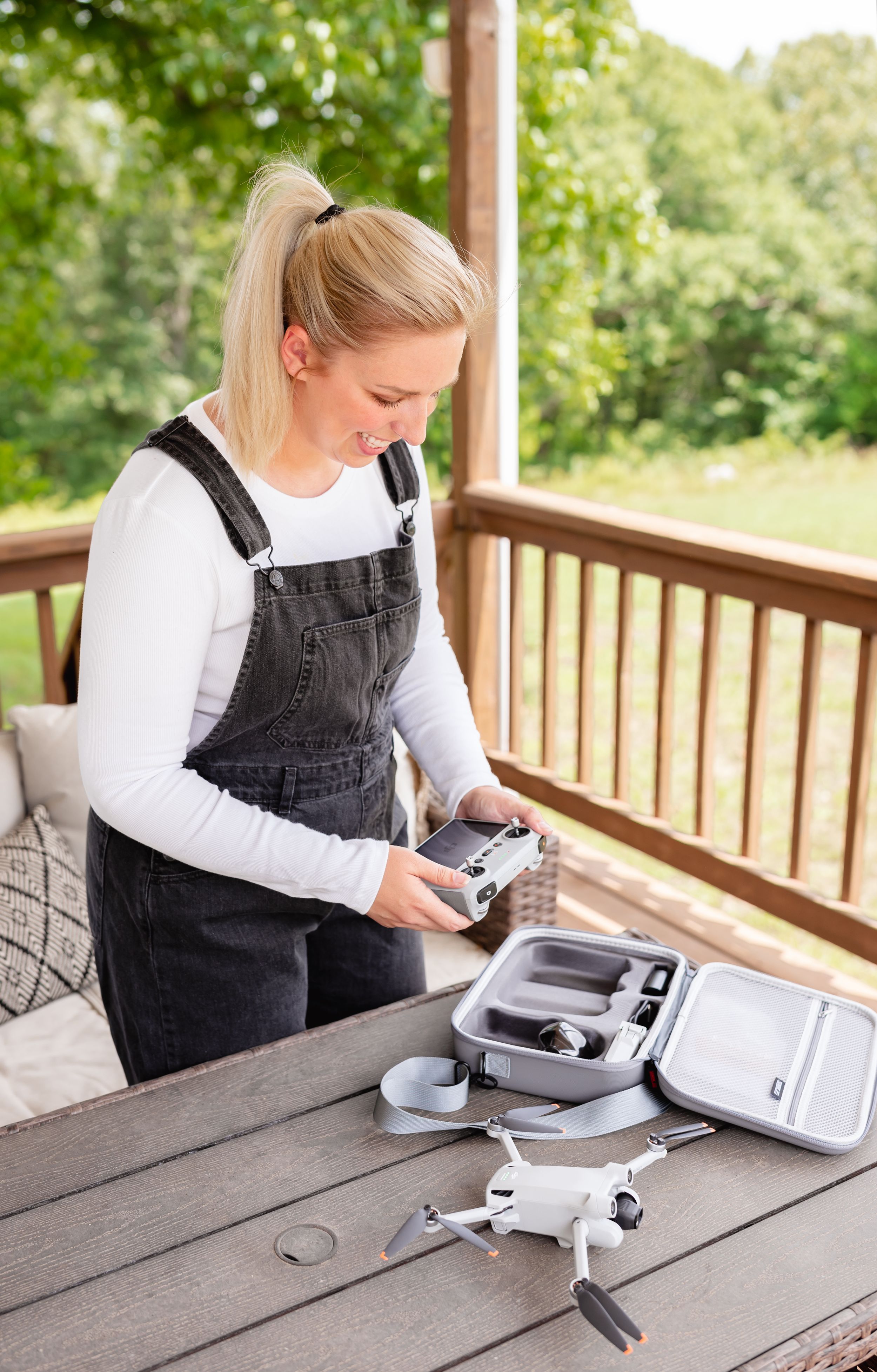 Blonde woman in denim overalls unpacking a DJI drone from its case on an outdoor wooden deck.