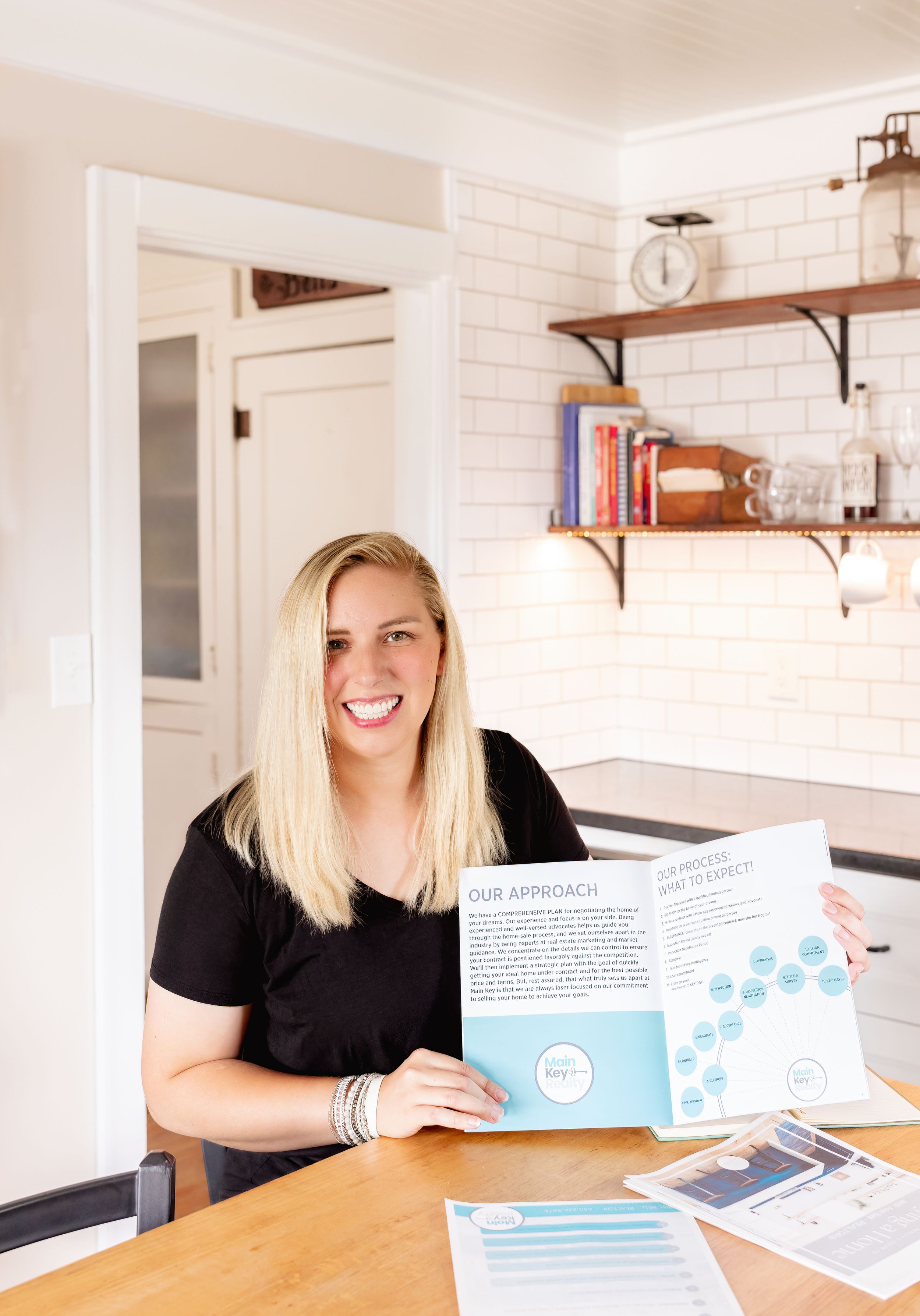 Smiling blonde woman holding a blue marketing brochure at a kitchen table with white subway tile backsplash.