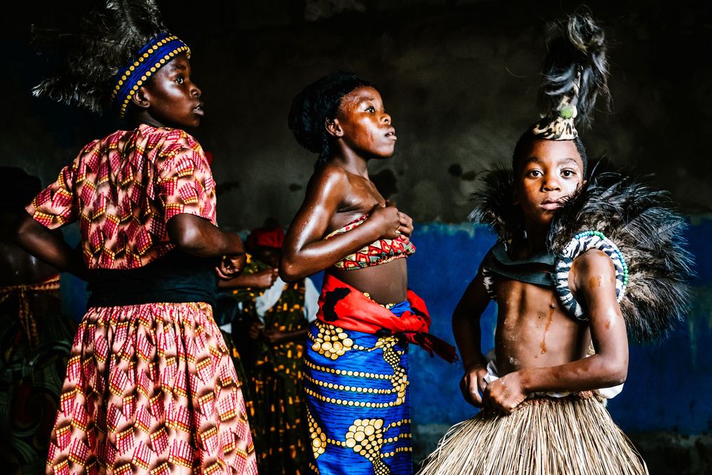 Three African women in traditional colorful attire and feathered accessories perform a cultural dance ceremony.