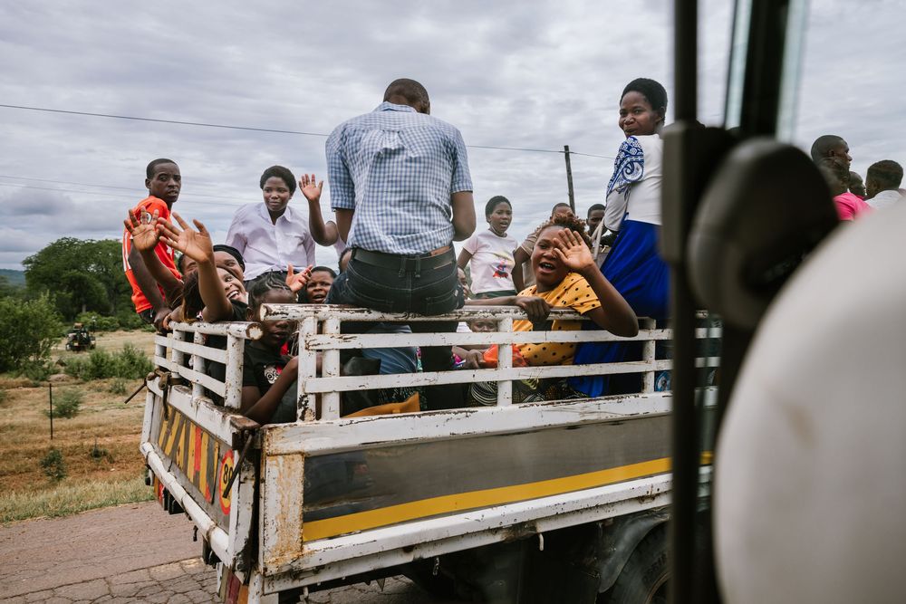 Group of people riding in the back of a truck on a rural road in Africa, waving and smiling at the camera.