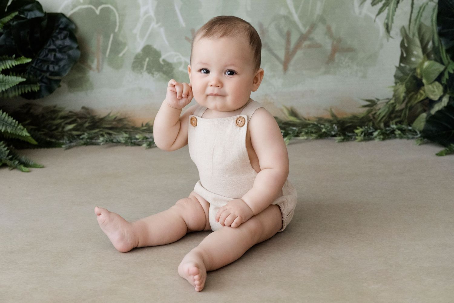 Adorable baby in beige romper sits on floor surrounded by lush greenery backdrop in professional photo.