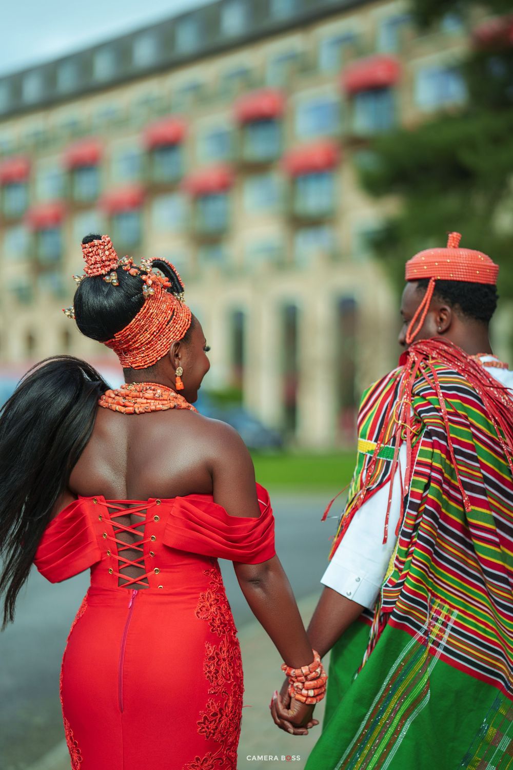 Nigerian couple in traditional Igbo attire holding hands, woman in red off-shoulder dress, man in colorful kente cloth.