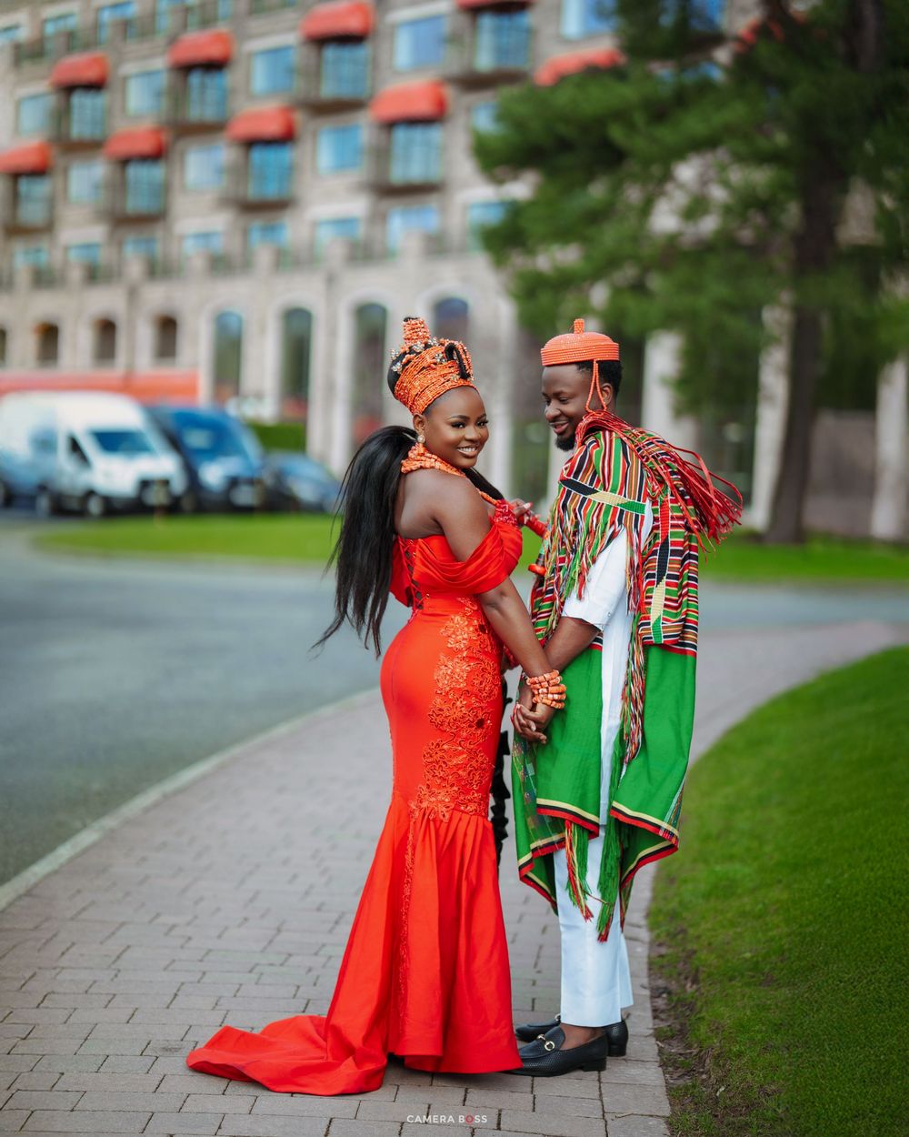 Couple in vibrant African traditional attire, woman in red mermaid gown, man in green and red outfit, posing outdoors.