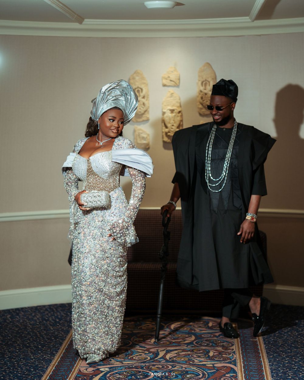 Couple in elegant Nigerian traditional attire, woman in silver gele and lace dress, man in black agbada with beaded necklace.