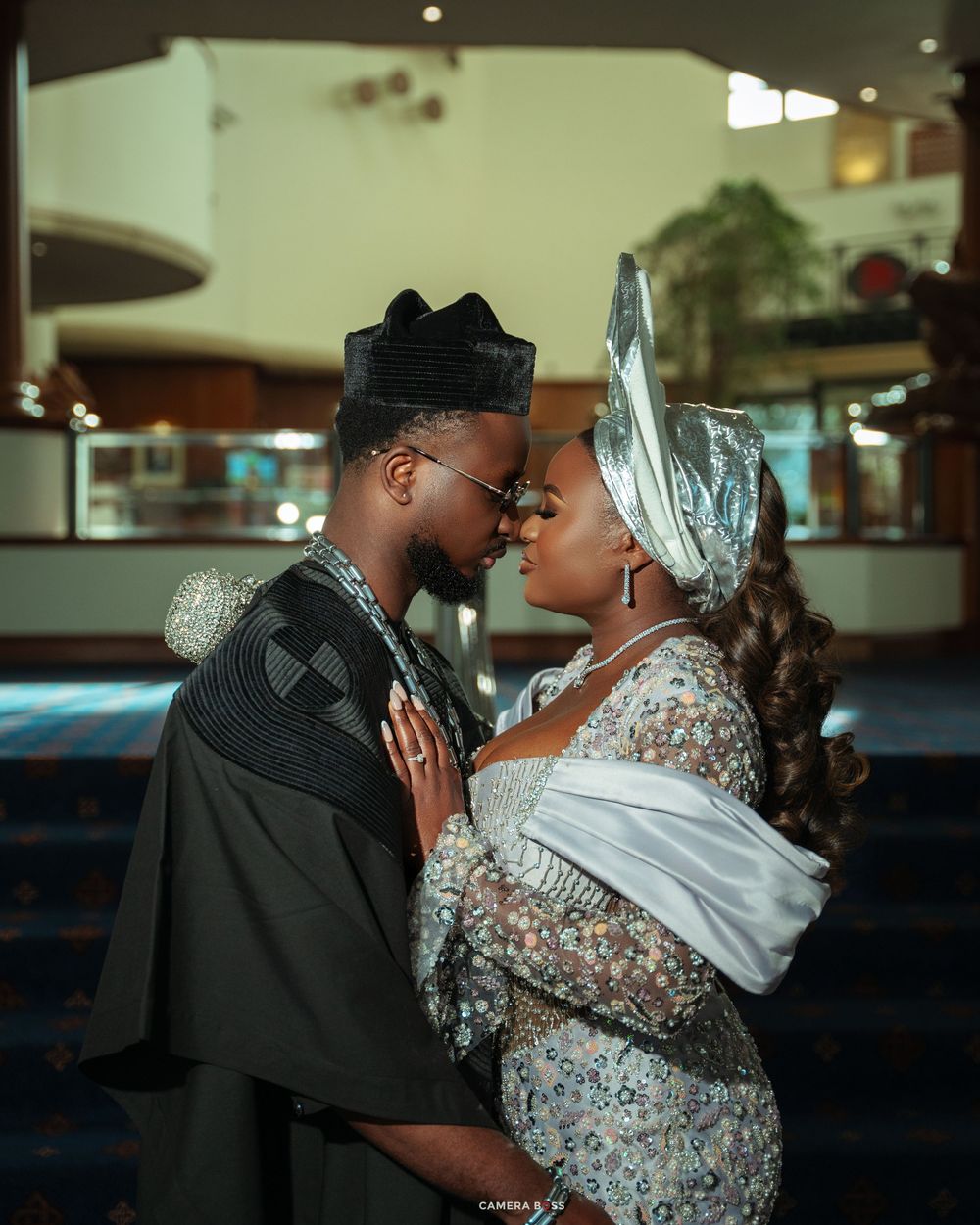 Couple in elegant Nigerian traditional attire sharing a romantic moment, man in black agbada, woman in silver gele headwrap.
