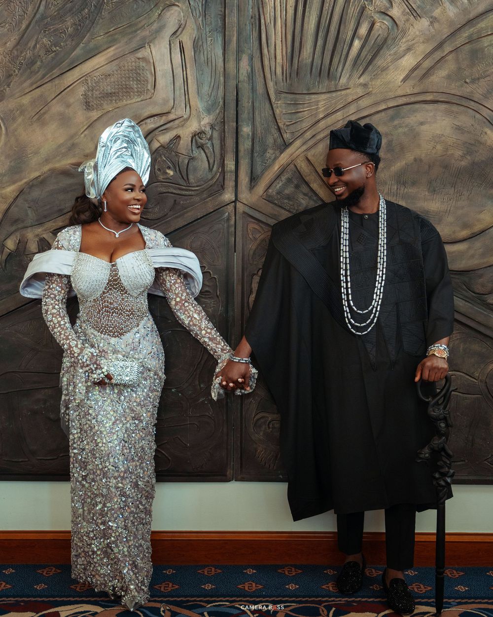 Couple in elegant Nigerian traditional attire, woman in silver gele and beaded gown, man in black agbada with pearl necklace.