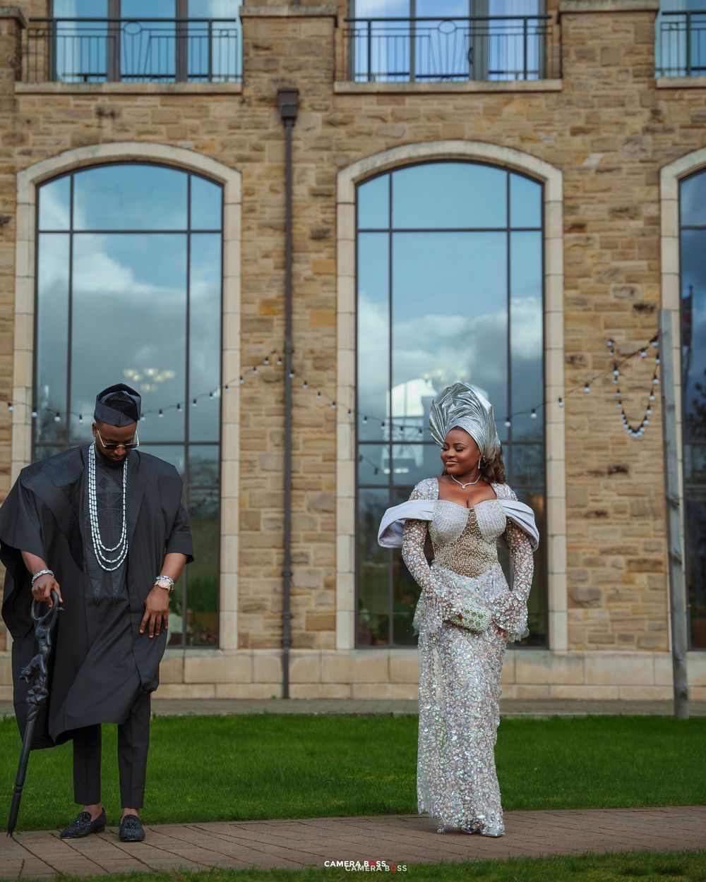 Nigerian couple in traditional attire, man in black agbada, woman in silver sequin gown with gele headwrap, outdoors.
