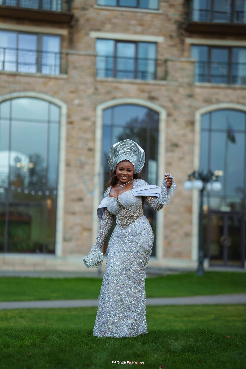 Woman in silver sequin gown and matching gele headwrap smiling outdoors at elegant stone building.
