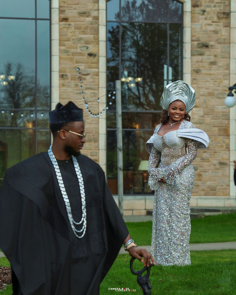 Nigerian couple in traditional Yoruba attire, man in black agbada, woman in silver sequin gown with gele headwrap.