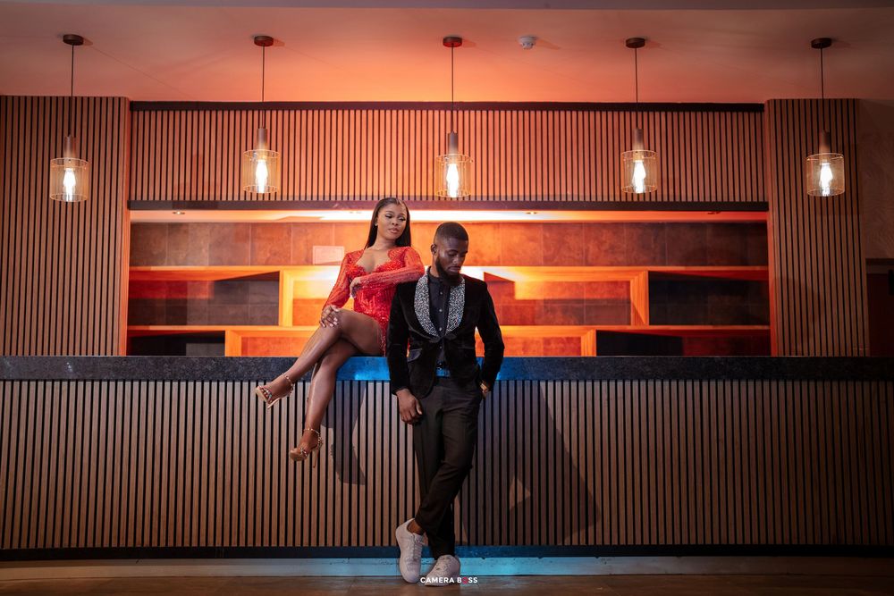 Couple posing elegantly in a modern bar, woman in red dress seated on counter, man in suit standing beside her.