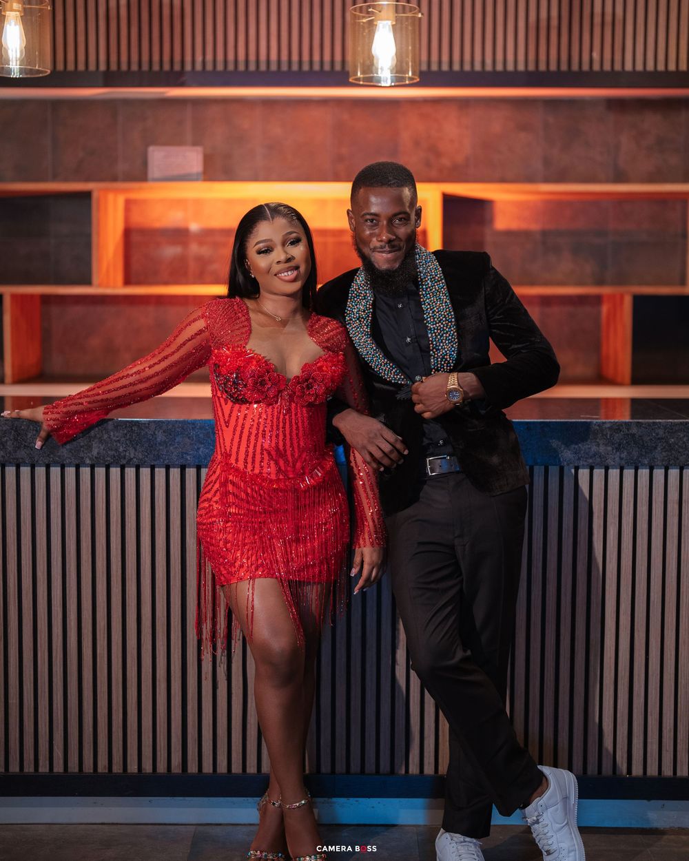 A couple poses elegantly, woman in red fringe dress and man in black suit with patterned lapel, at a stylish bar.
