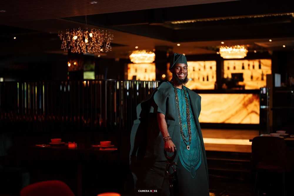 Man in traditional African green attire and cap poses confidently in a dimly lit elegant bar setting.