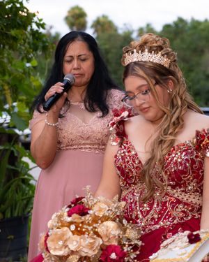 A young woman in a red quinceañera gown and tiara holds flowers beside a woman in pink speaking into a microphone.