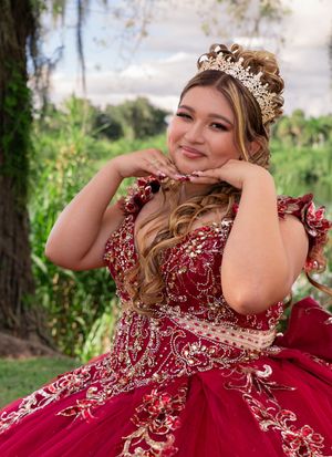 Young woman in red and gold quinceañera gown and tiara poses gracefully outdoors with lush green trees behind her.