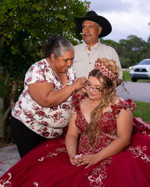 A young woman in a red quinceañera gown and crown is attended by her parents outdoors at dusk.