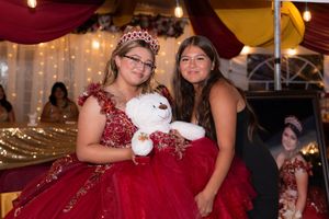 Two girls in red quinceañera dresses pose together holding a white teddy bear at a festive celebration.