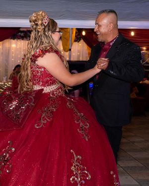 A young woman in a red quinceañera gown dances with a man in a black suit at a festive celebration.