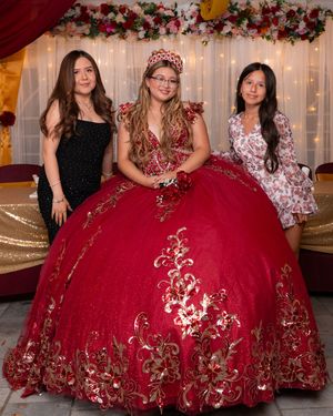 Girl in red gold quinceañera ball gown with crown poses with two friends at decorated celebration.