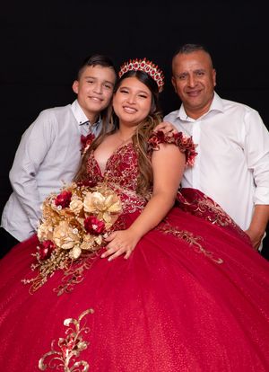 Girl in red quinceañera gown with tiara and bouquet poses with two male family members against black background.