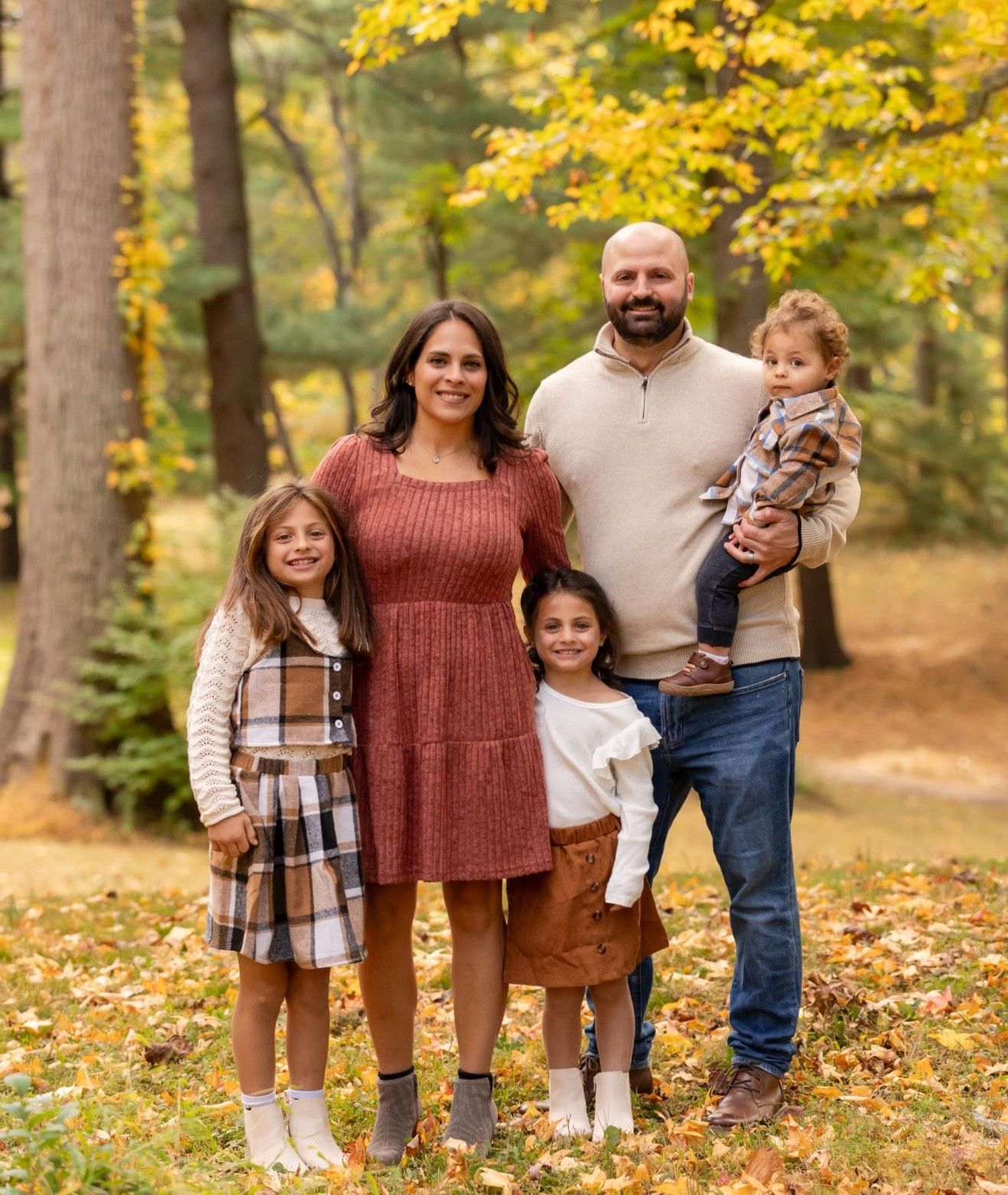 Family of five posing outdoors in autumn park with colorful fall foliage, wearing coordinated earth-tone outfits.