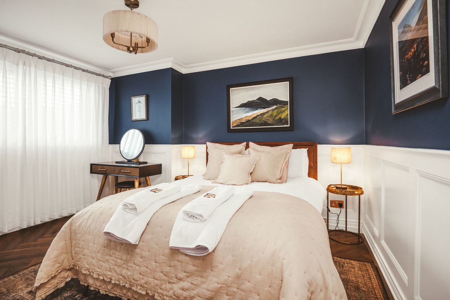 Elegant bedroom with navy blue walls, white wainscoting, beige bedding, and dark hardwood floors with landscape artwork.