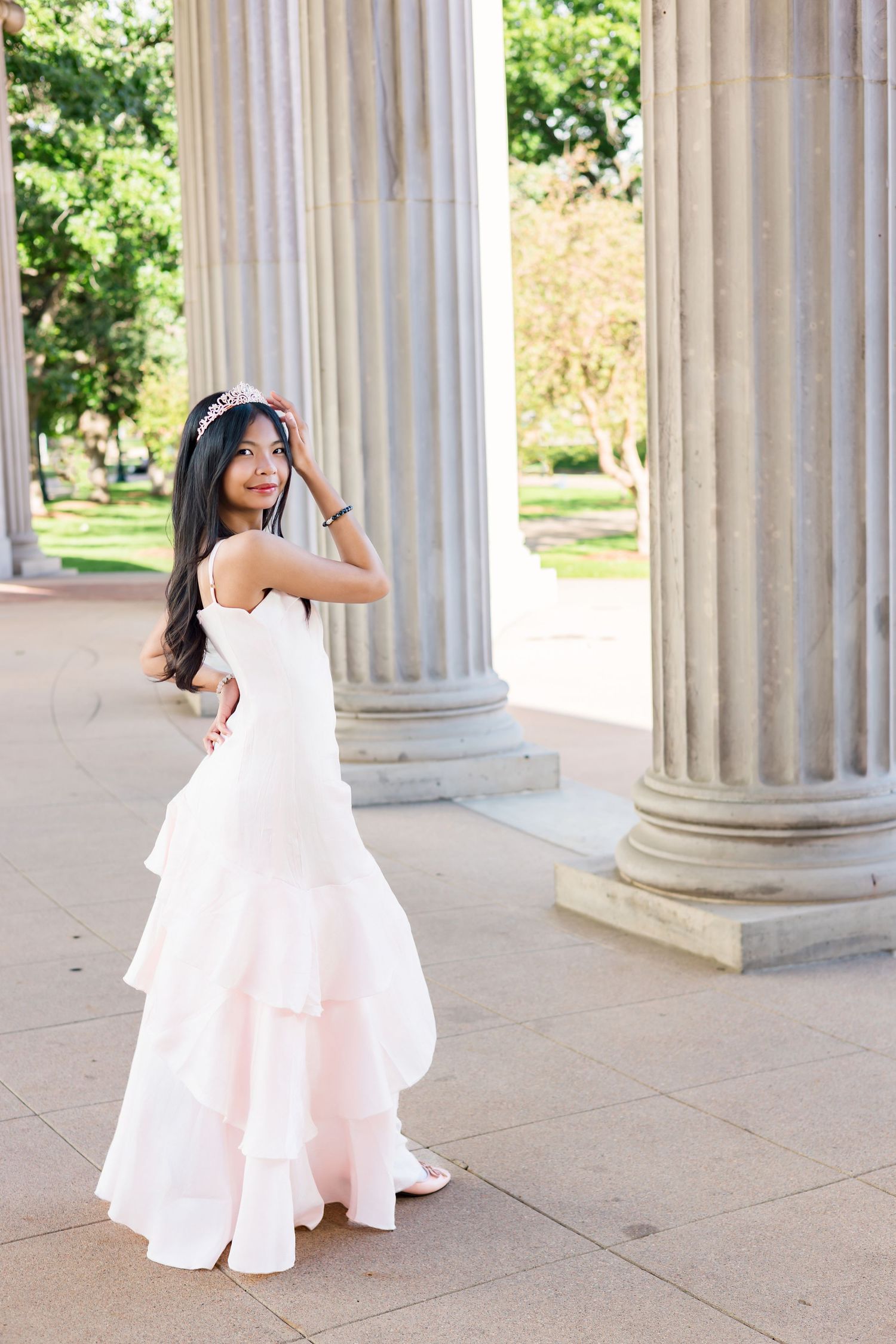 Young woman in white ruffled gown and tiara poses gracefully between classical stone columns in an outdoor setting.