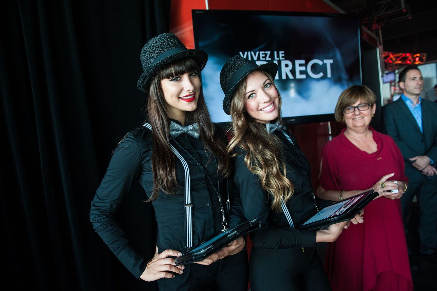 Two women in black outfits and hats pose at a Vivez Le Direct event with a guest holding a tablet.