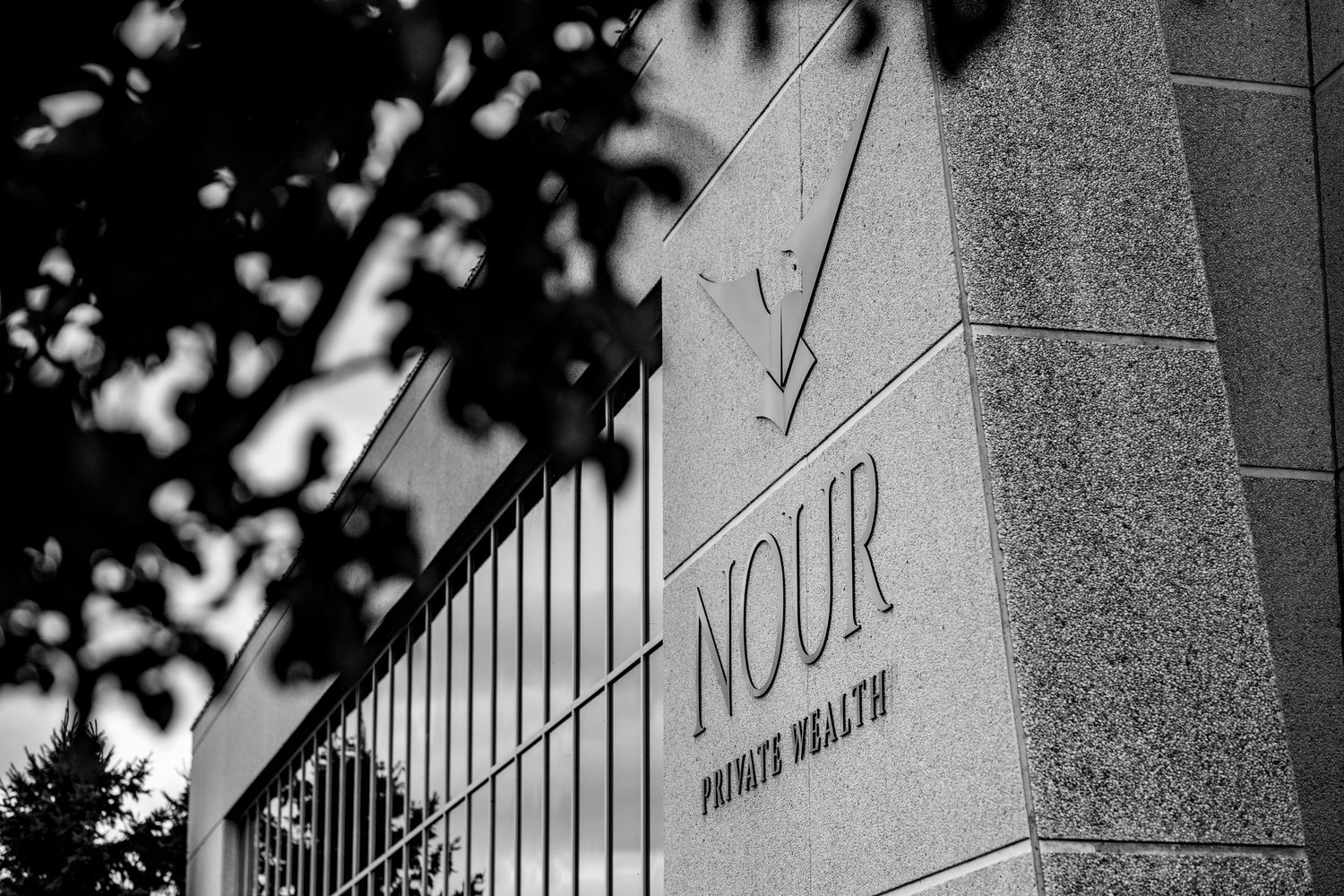 Black and white photo of Nour Private Wealth stone sign with decorative metal fence and trees in background.