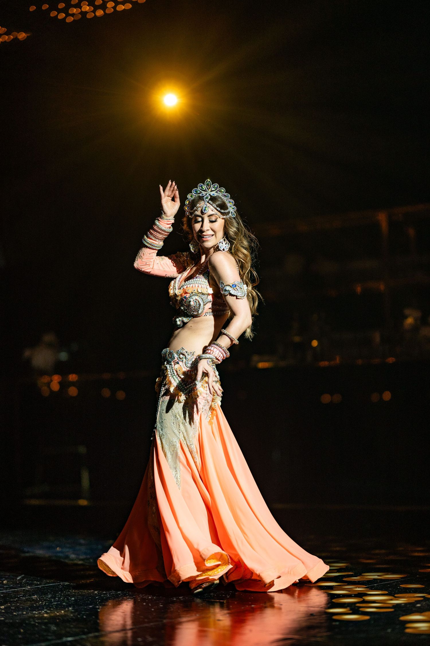 Belly dancer in orange costume and crown performs on dark stage under warm spotlight.