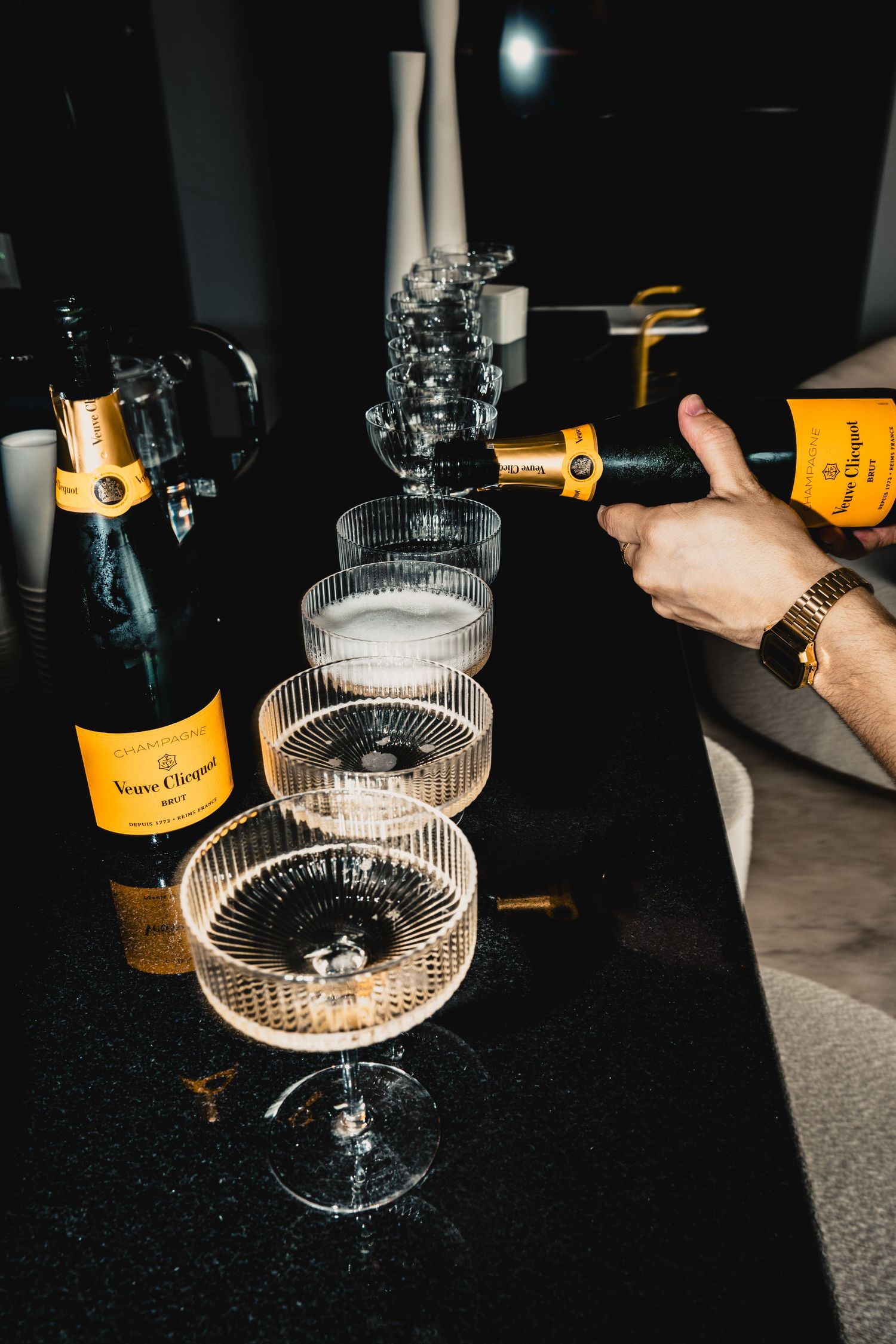 Hand pouring Veuve Clicquot champagne into stacked coupe glasses on a dark marble surface.